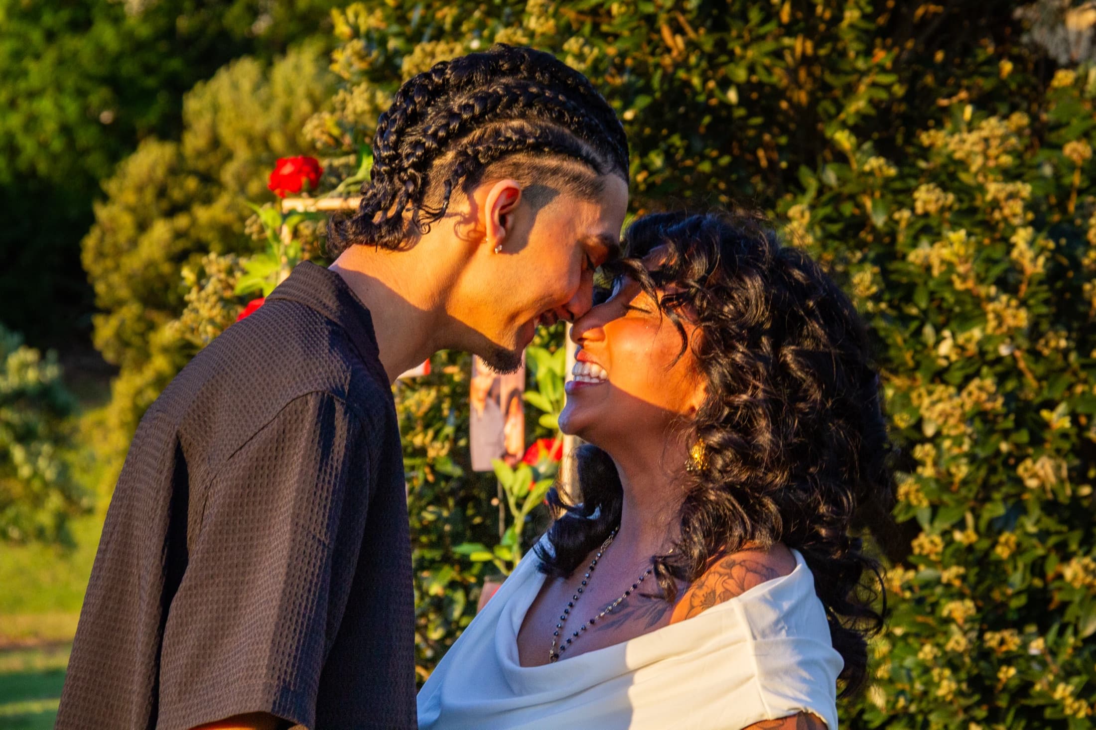 Dash and Robyn resting their foreheads together at golden hour, Robyn laughing, a rose bush behind them