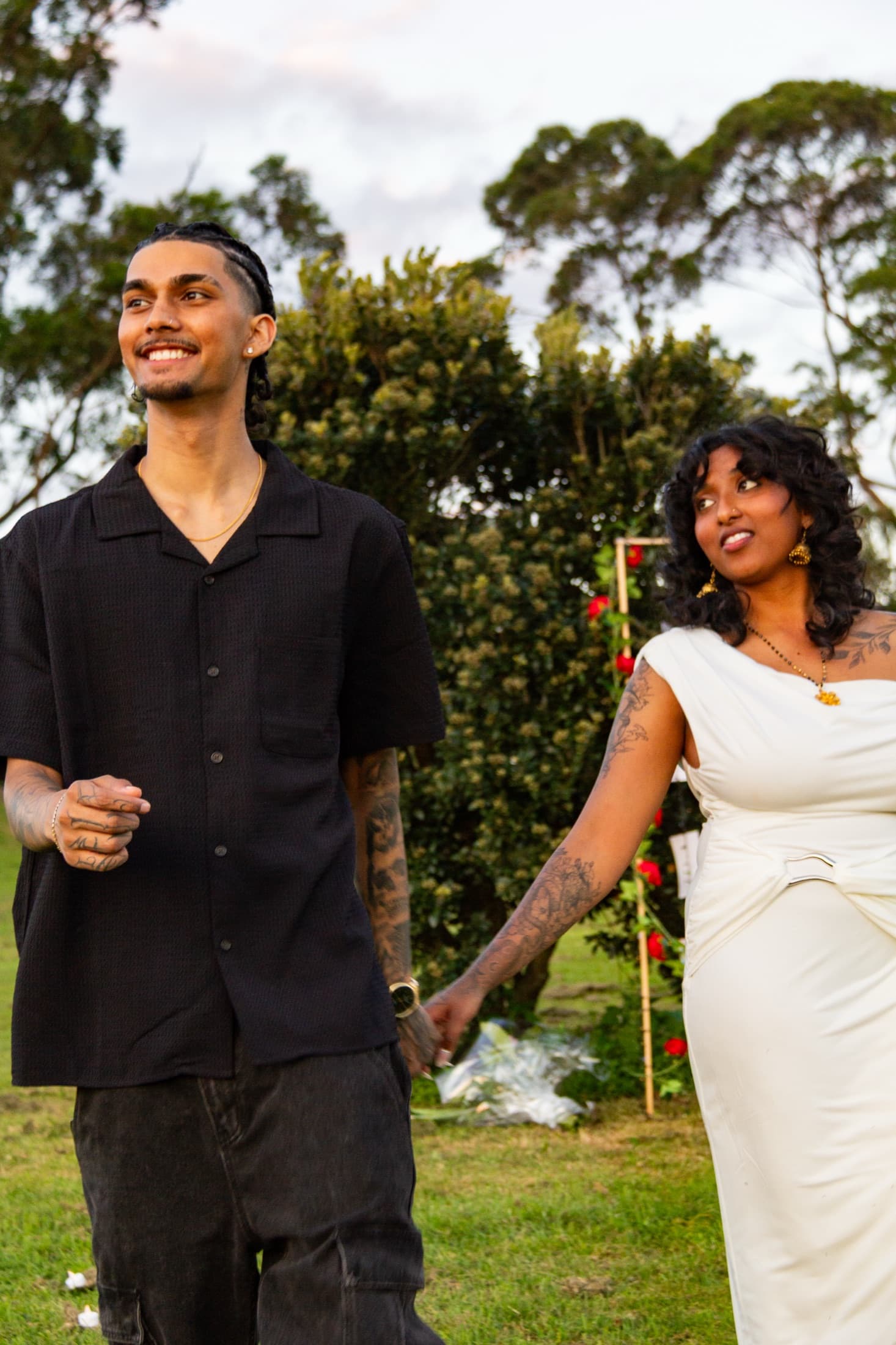 Dash and Robyn walking hand in hand across the grass, both smiling, roses and trees behind them