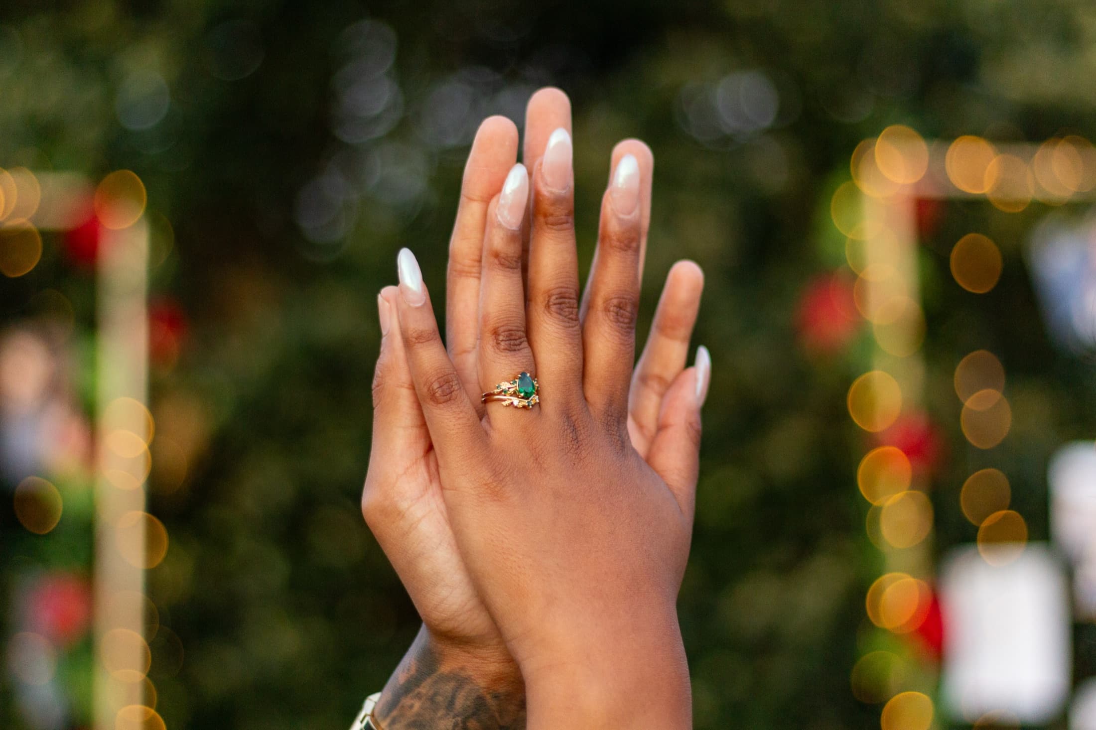 Robyn's hand raised to show her emerald engagement ring against warm fairy-light bokeh