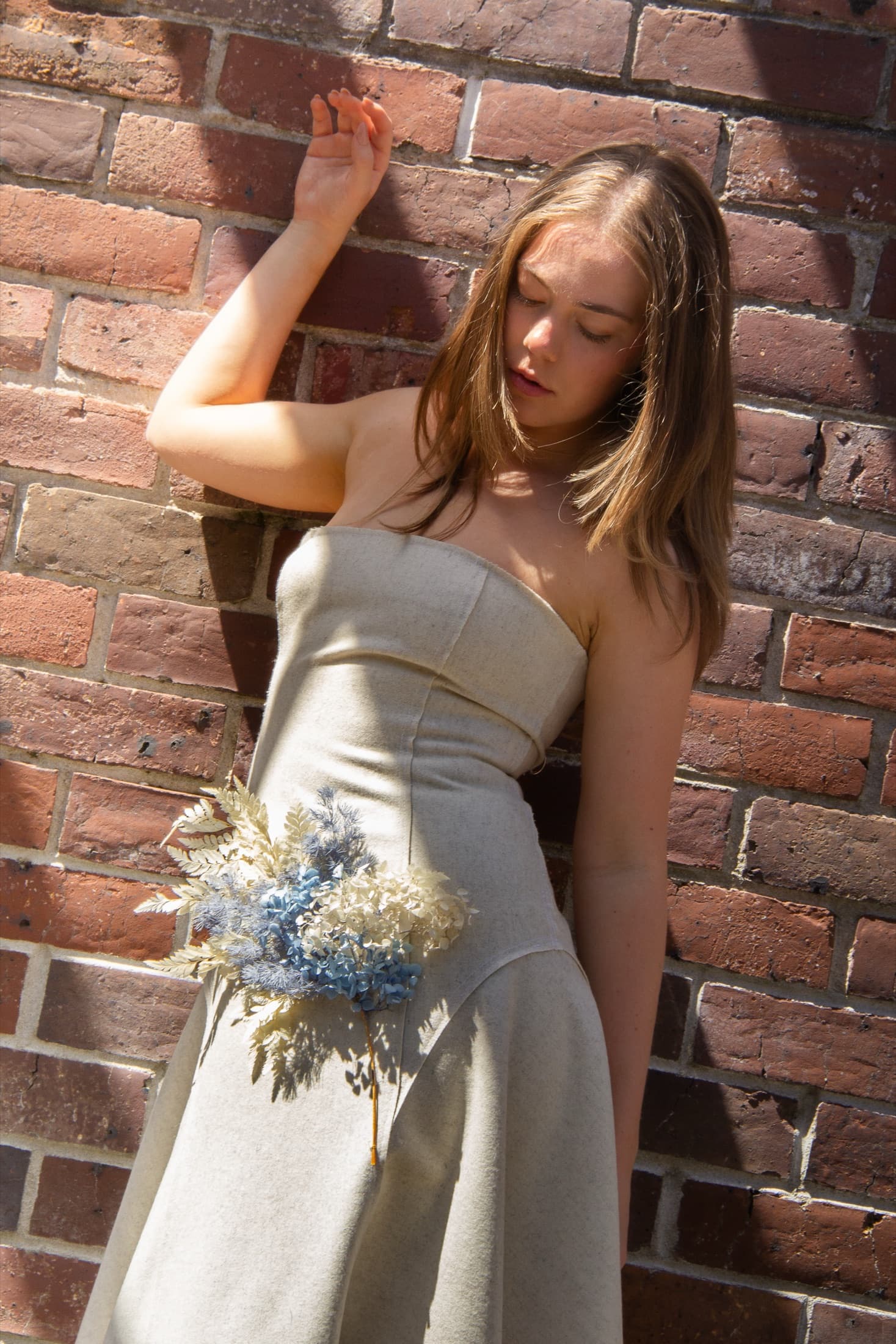 Model against a brick wall in a cream strapless dress embellished with blue dried flowers
