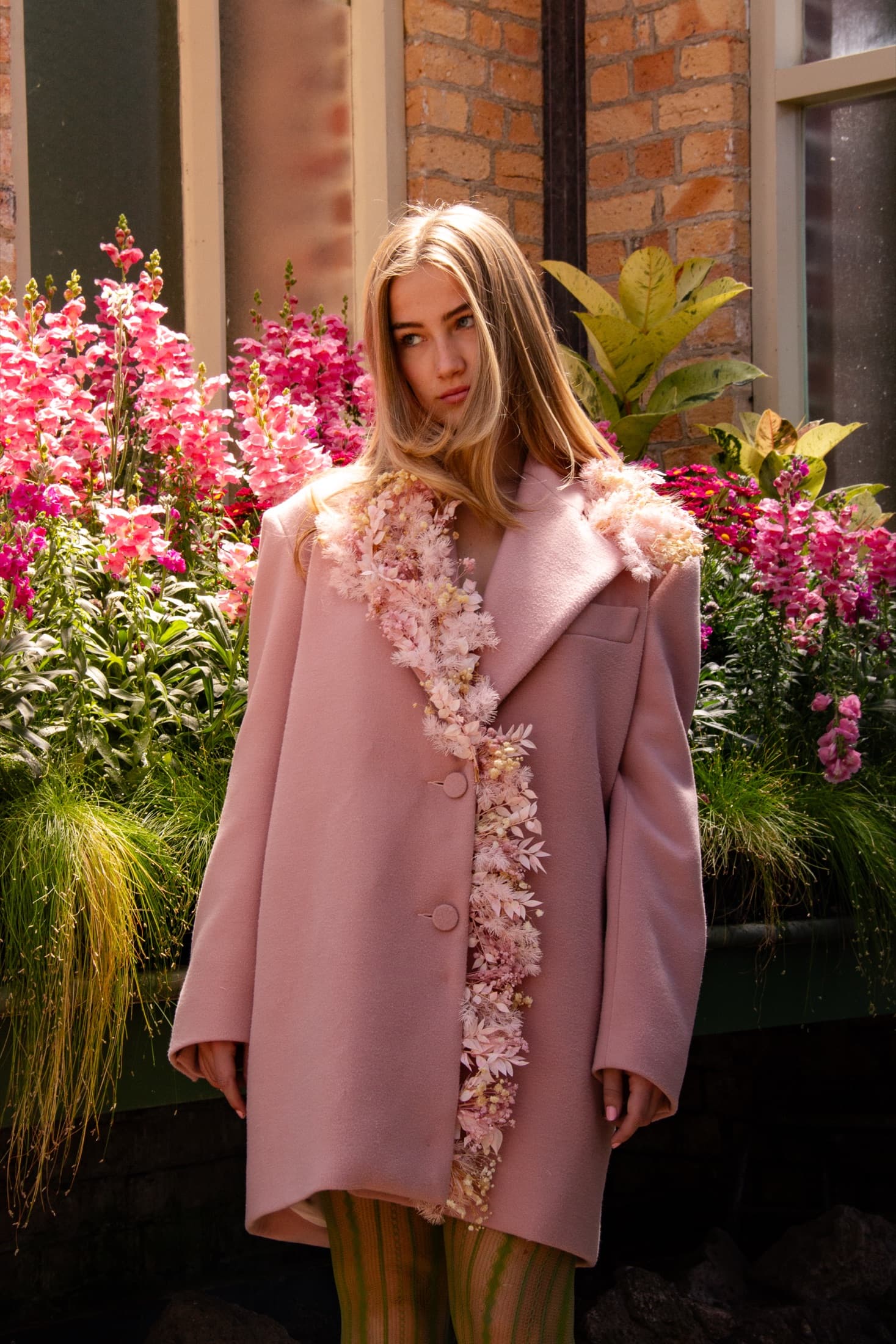 Model in a pink coat trimmed with dried flowers, standing among pink snapdragons
