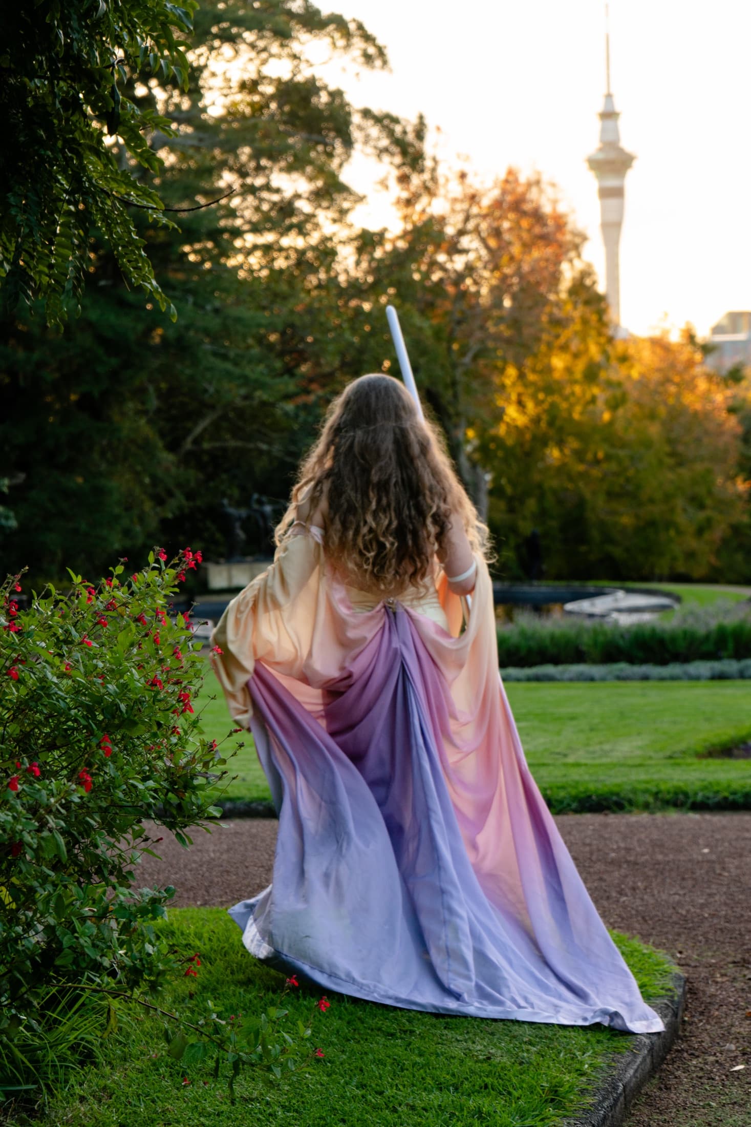 Model in a pastel gown holding an illuminated lightsaber in a lavender garden, Sky Tower behind at sunset